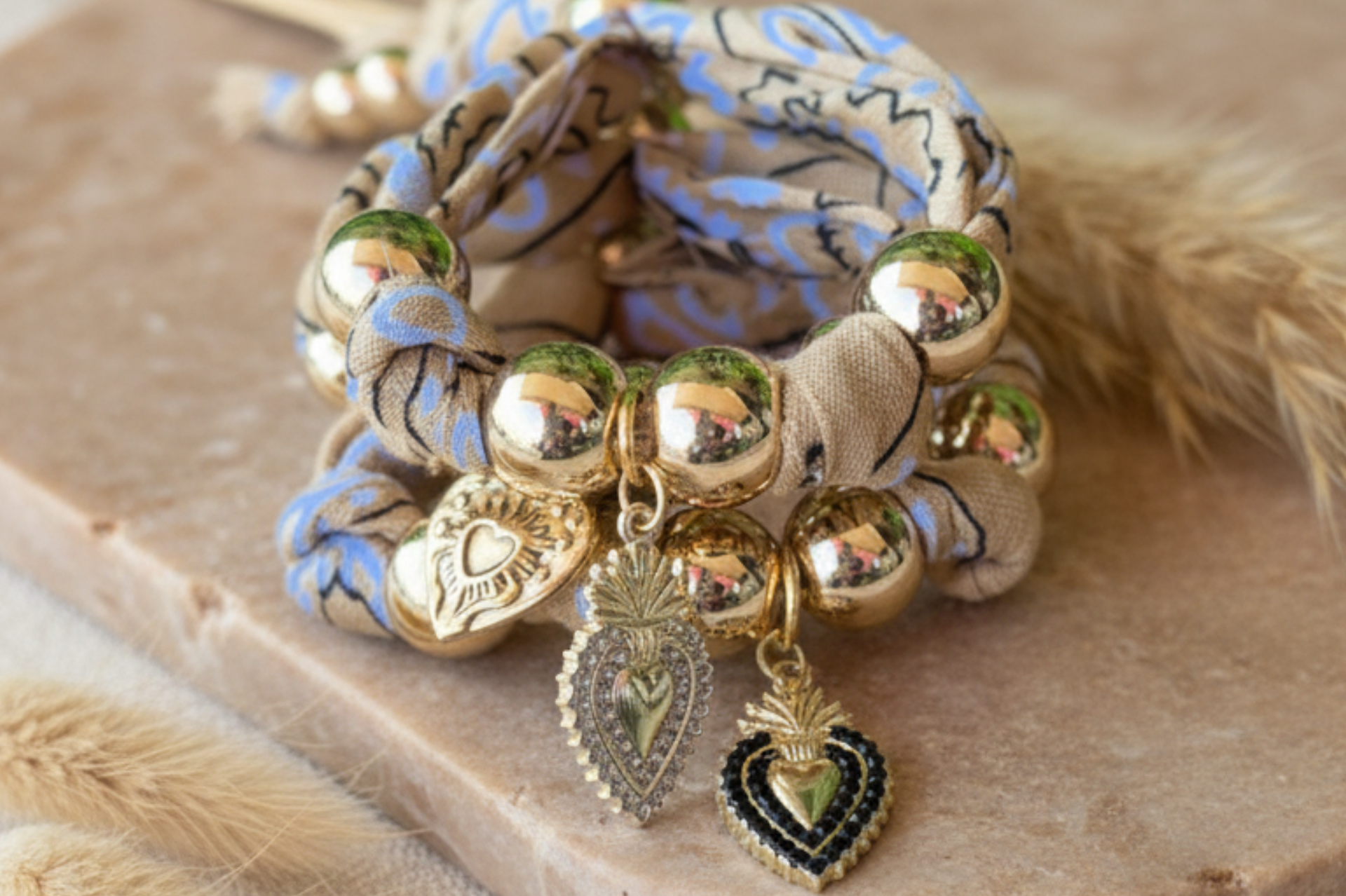 Gros plan sur un ensemble de bracelets en tissu bleu et blanc à motifs, agrémentés de grosses perles dorées et de charms en forme de cœurs sacrés argentés et noirs.