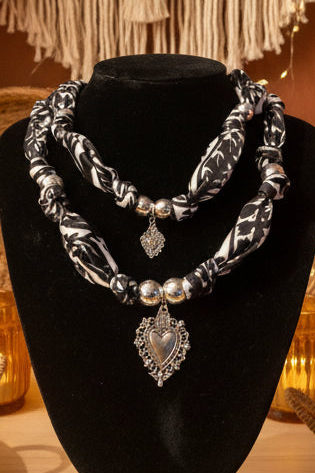 Femme portant le collier double en tissu noir et blanc avec pendentifs cœurs argentés, chapeau beige et décor architectural terracotta.