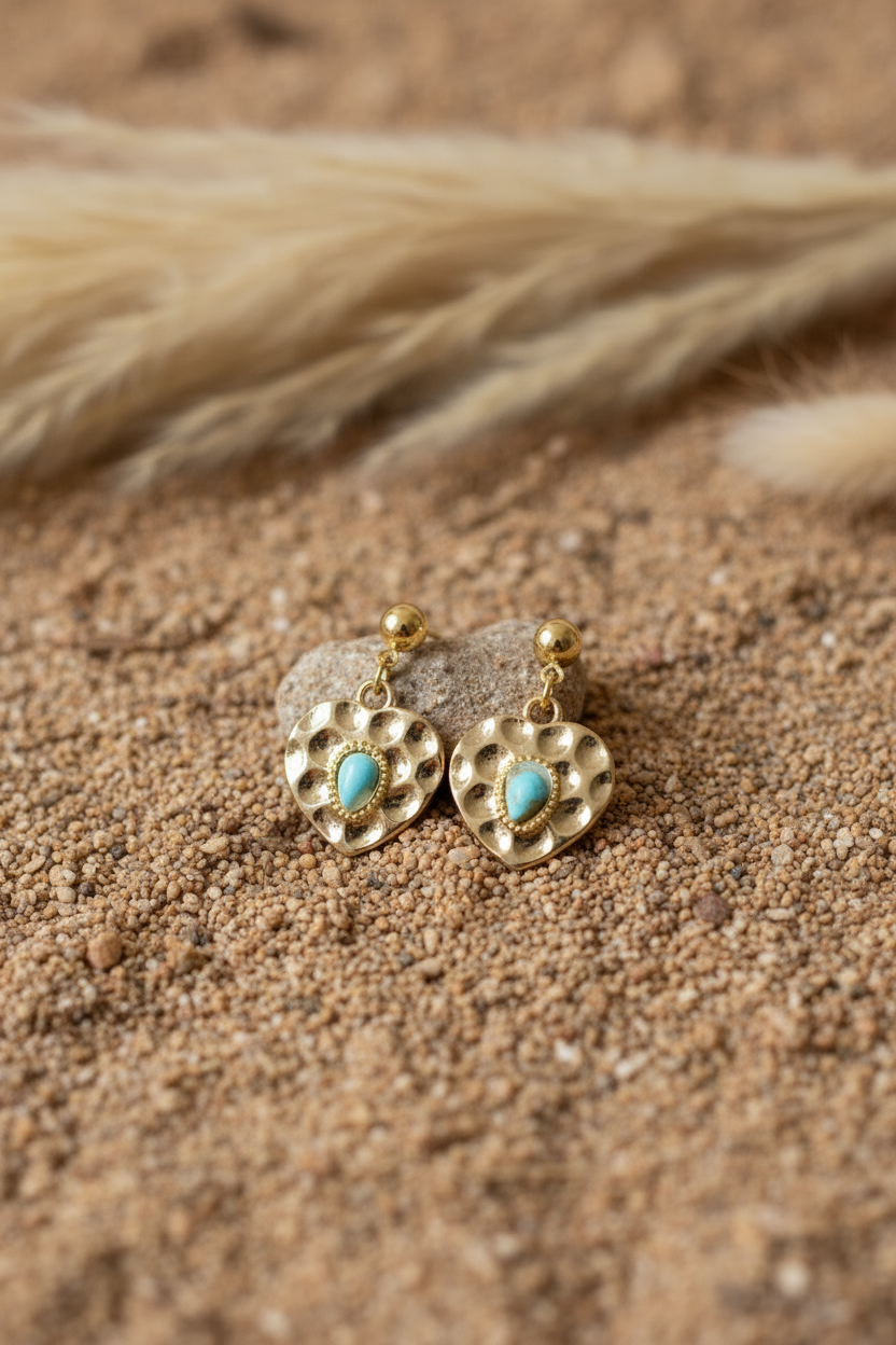 Détail des boucles d'oreilles cœurs dorés et perles turquoises Joyas by Jo, posées sur du sable chaud avec une touche de fleurs séchées pour un style bohème chic.