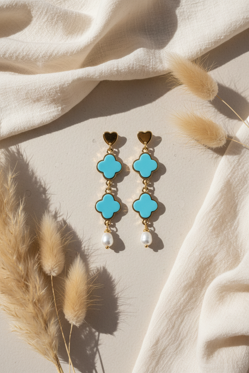 Paire de boucles d'oreilles pendantes Joyas by Jo avec trèfles bleu turquoise, coeurs dorés et perles d'eau douce sur un fond de lin beige et fleurs séchées.