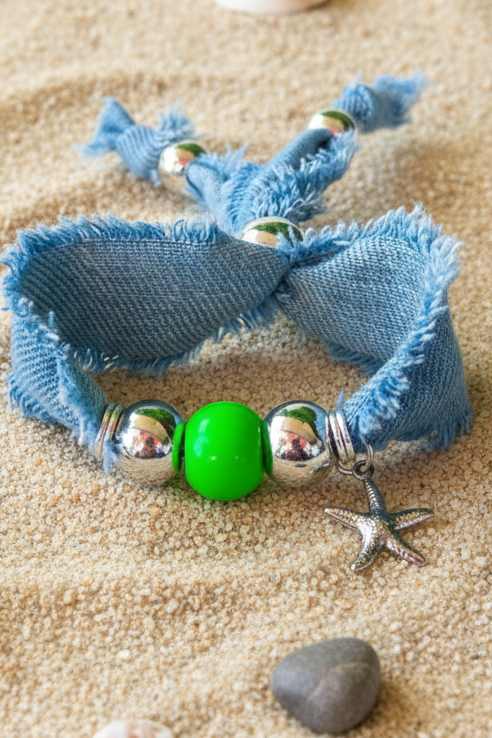 Vue détaillée du bracelet "Escale Denim" en tissu bleu jean, orné d'une perle verte centrale, de perles argentées et de charms étoile de mer, posé sur du sable avec des petits galets. Joyas by jo