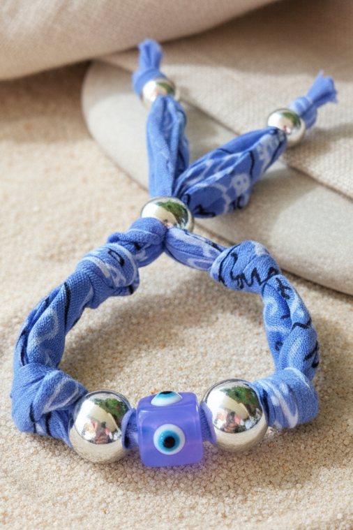 Détail du bracelet Anita fabriqué en tissu bandana bleu tressé avec une perle centrale Nazar (œil protecteur) et des perles argentées, posé sur du sable fin. Joyas by jo