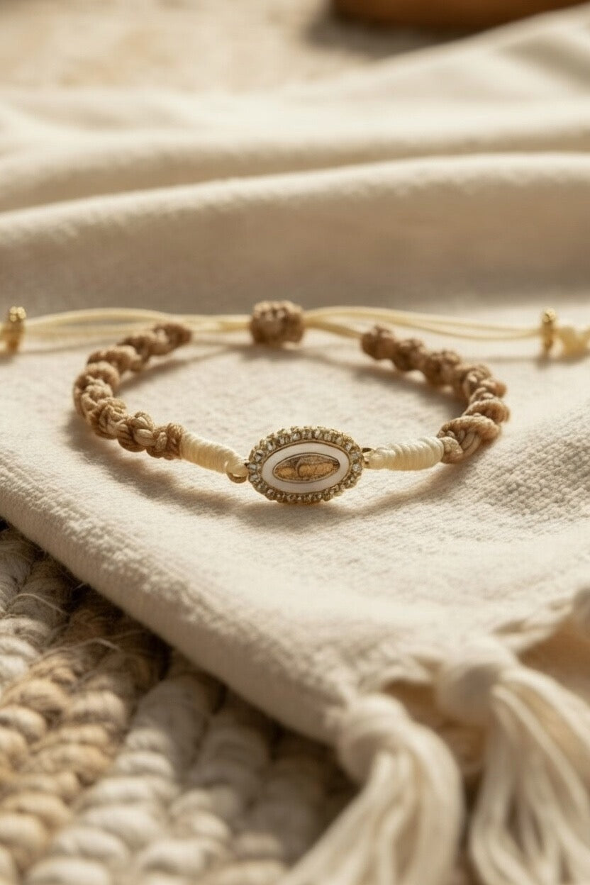 Bracelet Joyas by Jo en cordon tressé beige et sable avec médaillon ovale doré orné de mains en prière et cristaux brillants posé sur un tissu en lin naturel.