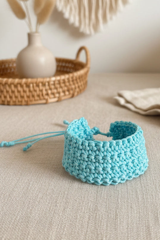 Bracelet large en maille crochetée bleu turquoise posé sur une nappe en lin beige à côté d'un panier en osier.