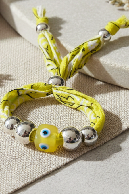 Vue rapprochée du bracelet Elena en tissu bandana jaune tressé, orné de perles argentées et d'une perle centrale jaune "œil protecteur", posé sur un support minéral. Joyas by jo
