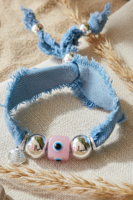 Vue de dessus d'un bracelet artisanal en tissu denim bleu clair effiloché, posé sur une pochette en lin beige. Le bijou est orné de deux grandes perles sphériques argentées encadrant une perle centrale rectangulaire rose portant un motif d'œil porte-bonheur. Un petit charm argenté en forme de coquillage pend sur le côté. Le décor en arrière-plan se compose de sable de plage fin, de galets arrondis et de quelques brins d'herbe séchée.