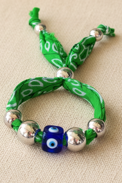 Gros plan du bracelet Julia "Regard d'Azur" montrant un cordon tressé vert, des perles argentées et une perle centrale œil bleu (nazar), posé sur un tissu neutre. Joyas by jo