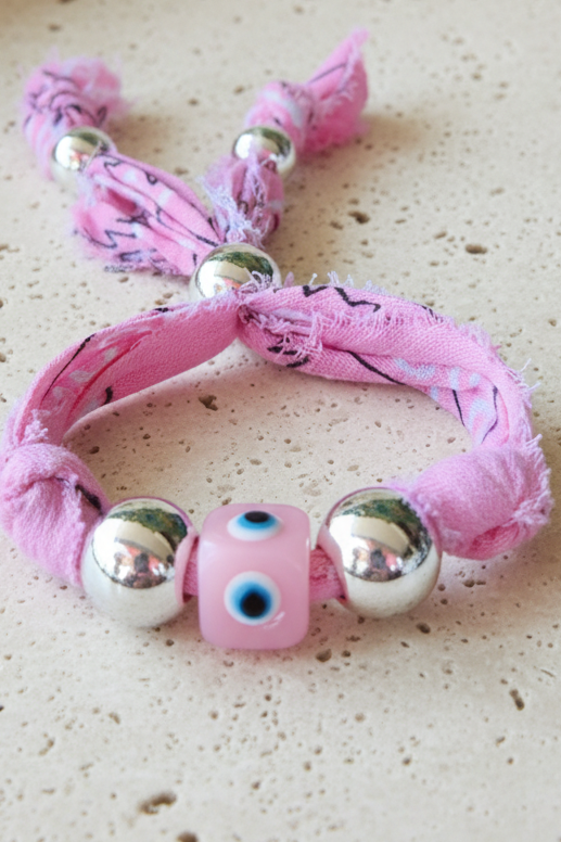 Gros plan du bracelet Ludmilla en tissu rose fleuri, mettant en relief sa perle centrale "œil" rose et ses perles argentées sur un fond clair épuré. Joyas by jo