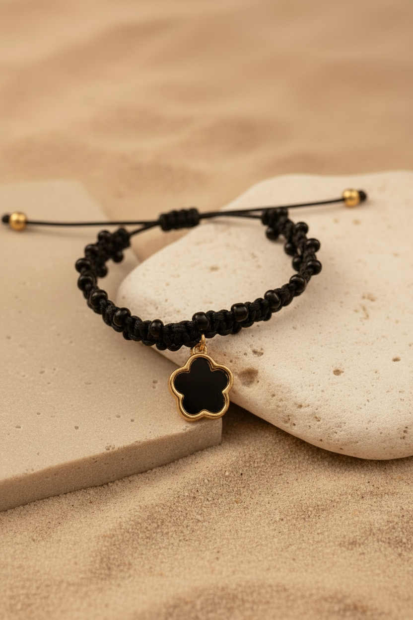 Bracelet artisanal en perles noires avec pendentif trèfle doré et noir, posé sur des pierres blanches et du sable fin, Joyas by Jo.