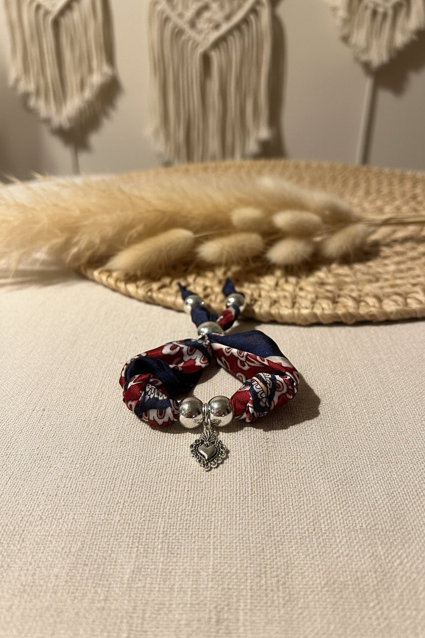 Bracelet en soie Paloma avec pendentif cœur argenté posé sur un support en osier naturel.