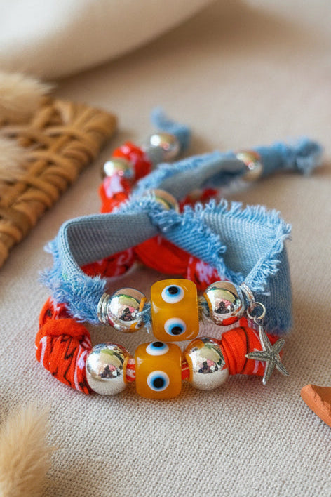 Gros plan sur deux bracelets en textile. L'un est en jean bleu clair effiloché avec une perle carrée jaune et un motif oeil bleu au centre. L'autre est en tissu rouge orangé. On distingue des perles argentées et un petit pendentif étoile de mer métallique.