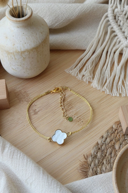 Bracelet minimaliste en fil doré résistant avec pendentif trèfle bleu turquoise posé sur un support décoratif blanc.