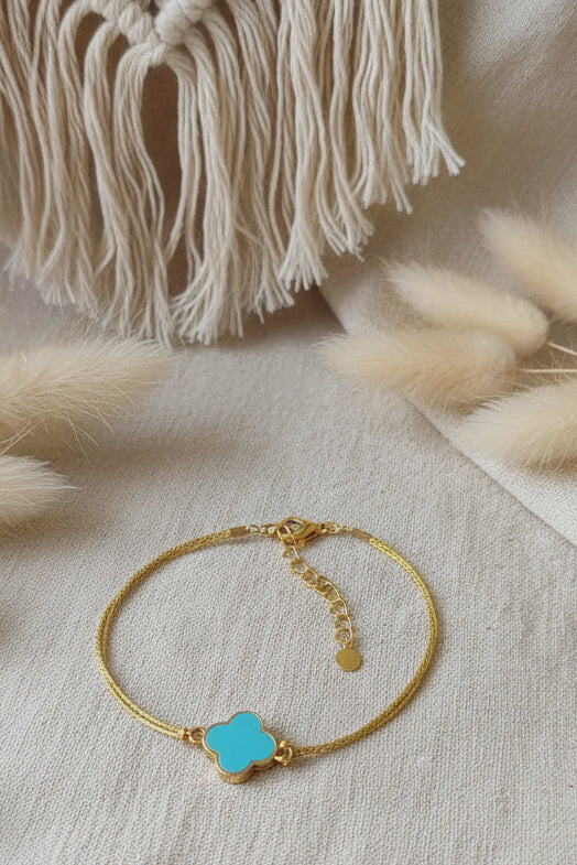 Bracelet minimaliste en fil doré résistant orné d'un pendentif trèfle bleu turquoise sur un fond de décoration bohème.