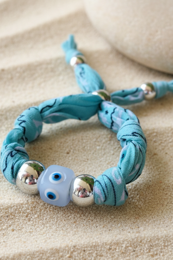 Gros plan du bracelet Véra "Amulette Turquoise" posé sur du sable blanc, mettant en avant son cordon en tissu bleu azur, ses perles argentées et sa perle centrale œil bleu clair. Joyas by jo