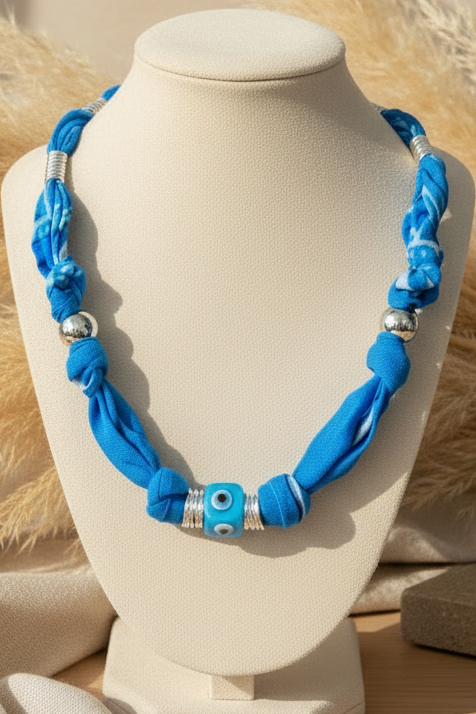 Collier Antonia "Azur Éternel" présenté sur un buste beige. Le bijou est composé d'un cordon en tissu bleu azur texturé, de perles argentées et d'une perle centrale "œil protecteur" bleue et blanche. Joyas by jo