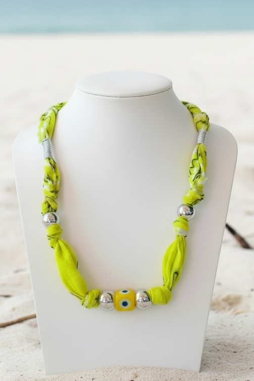 Collier Aylin "Œil Protecteur Citron" exposé sur un buste blanc posé sur le sable. Le bijou se compose d'un cordon jaune fluo éclatant, de perles argentées et d'une perle centrale "œil" en céramique jaune. Joyas by jo