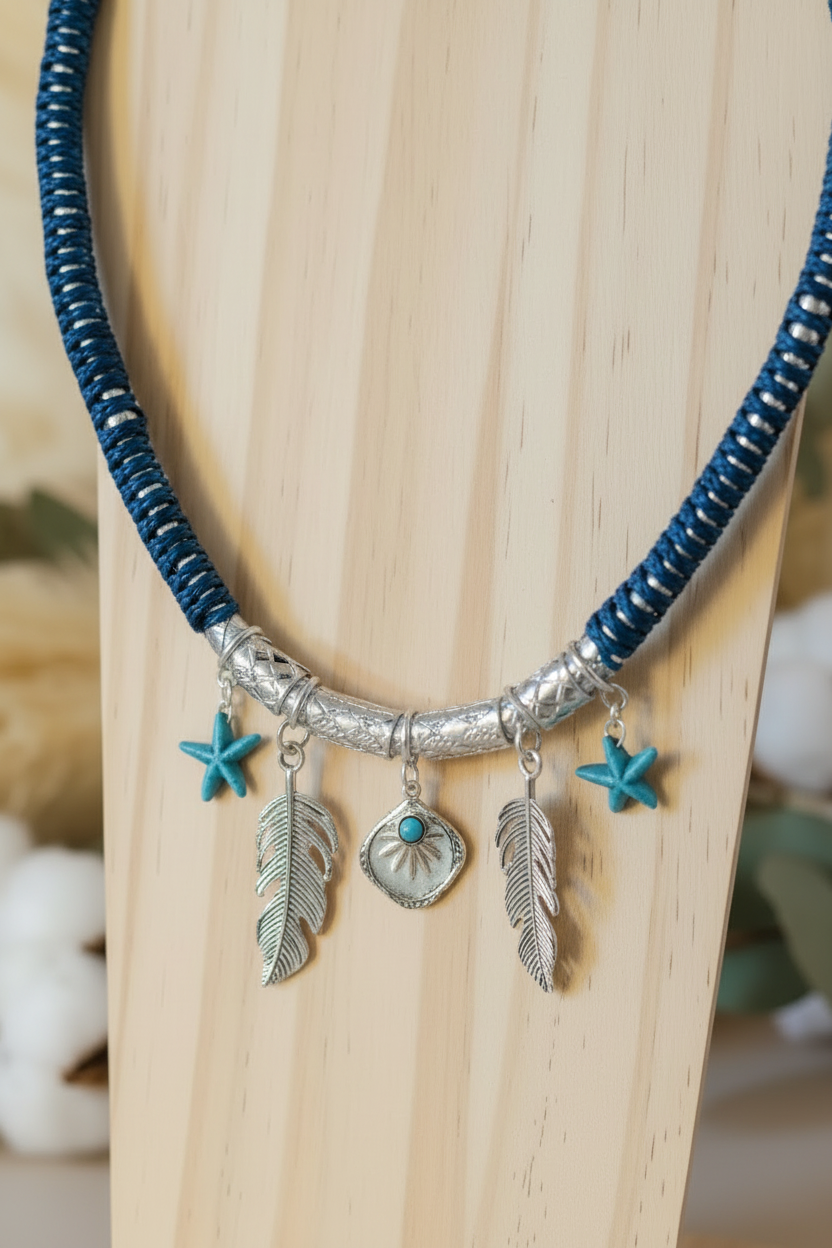 Gros plan d'un collier Joyas by Jo en cordon bleu tressé avec pendentifs plumes en argent et étoiles turquoises sur un support en bois.
