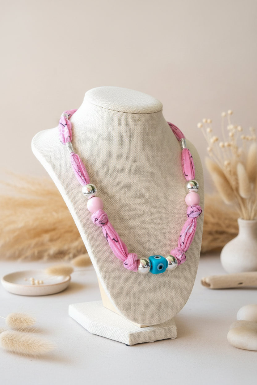 Collier Irina "Harmonie Pastel" exposé sur un buste blanc. Il est composé d'un cordon en tissu rose doux, de perles argentées et d'une perle centrale "Nazar" (œil protecteur) bleue et blanche. Joyas by jo