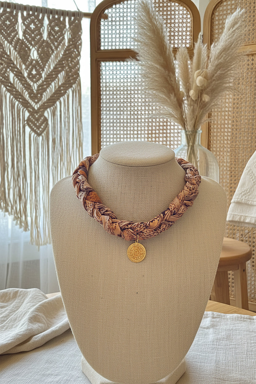Collier plastron tressé en tissu aux tons chauds avec un médaillon doré central, présenté sur un buste d'exposition blanc.