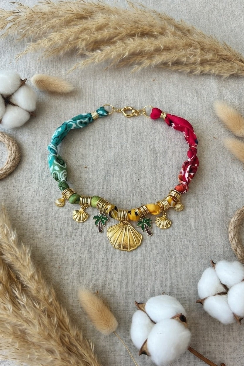 Collier artisanal Joyas by Jo en tissu multicolore avec pendentifs coquillages dorés et palmiers sur fond de lin naturel.