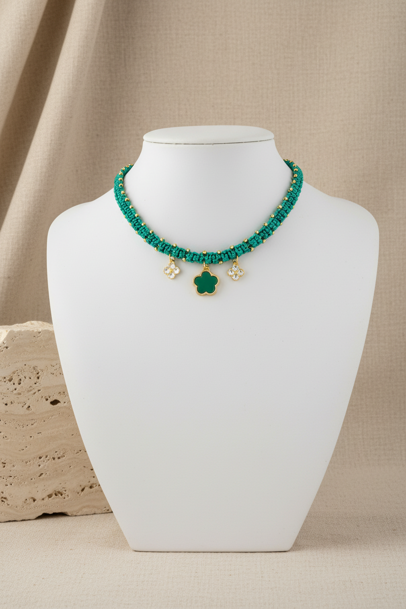 Zoom sur le collier tressé vert émeraude avec charms fleurs dorées Joyas by Jo sur un buste d'exposition blanc.