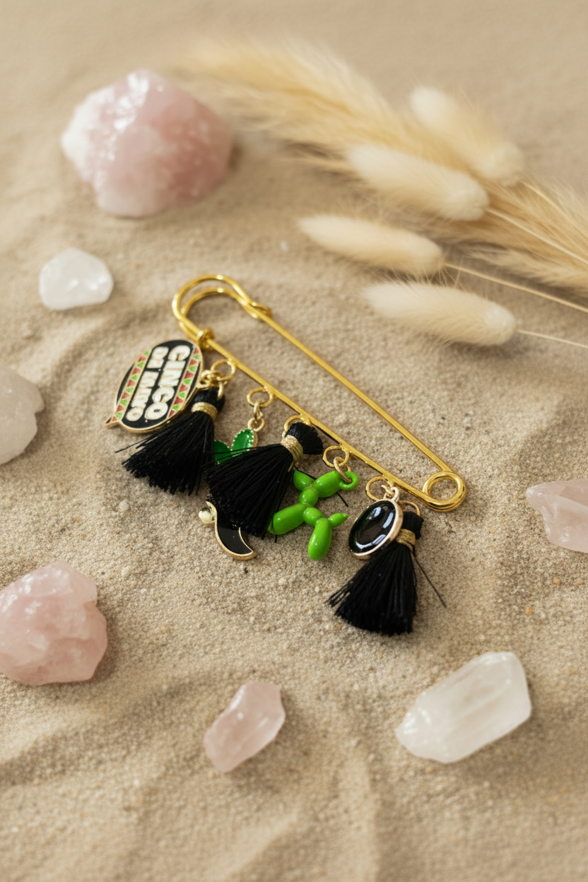 Épingle de sûreté dorée de style broche ornée de plusieurs breloques : un pompon noir, un cœur, un chien de baudruche vert, un smiley noir et un cactus vert. L'objet est disposé sur un tissu beige texturé, entouré de pierres naturelles, de cristaux de quartz et de fleurs séchées.