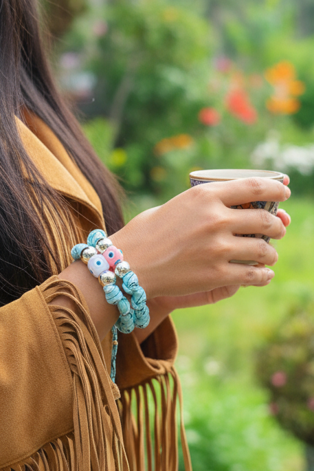 Gros plan sur le poignet d'une femme portant plusieurs bracelets Joyas by jo. On distingue les textures contrastées du denim bleu et des tissus rouges à motifs. Les détails incluent des perles oeil protecteur jaunes et des éléments argentés brillants.