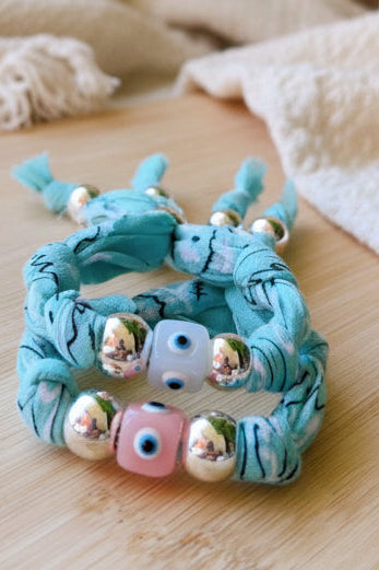 Bracelet Joyas by jo en denim bleu clair avec des bords effilochés, présenté sur fond blanc. Au centre, une perle jaune avec un motif oeil bleu est entourée de perles argentées. Une petite étoile de mer métallique est suspendue au tissu.