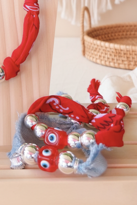 Bracelet de la marque Joyas by jo composé d'un tissu rouge à motifs et d'un liseré en denim bleu. Il est décoré d'une perle carrée jaune avec un oeil bleu, de billes argentées et d'une étoile de mer en métal. Le bijou est présenté à plat sur un fond blanc.