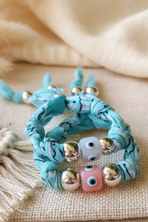 Un bracelet Joyas by jo posé à plat sur fond blanc. Il mélange un tissu rouge à motifs blancs et une bande de denim bleu clair. Le bijou est orné de billes argentées, d'une perle carrée jaune avec un oeil bleu et d'une breloque étoile de mer en métal.