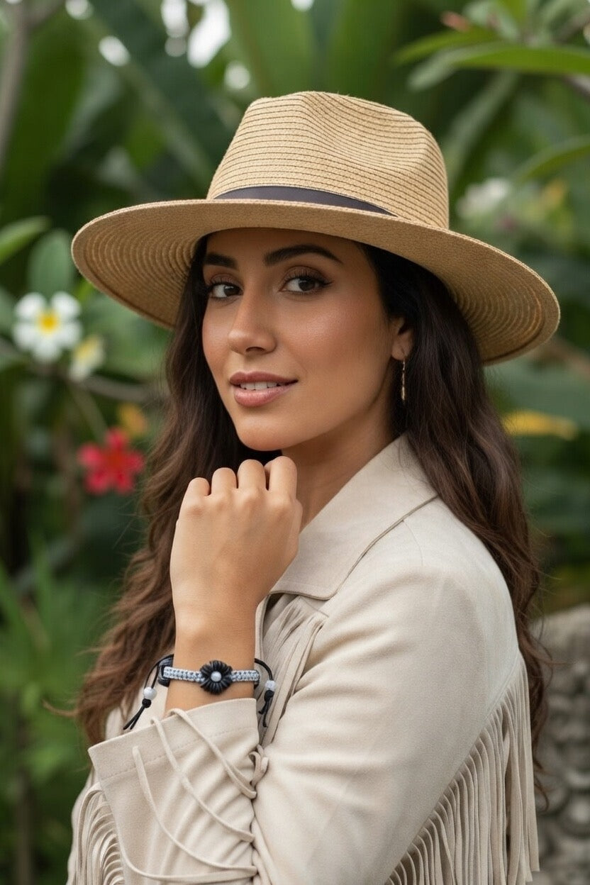 Une femme portant un chapeau de paille et une veste à franges beige met en avant un bracelet artisanal Joyas by Jo orné d’une fleur noire au centre, sur un fond de jardin tropical luxuriant.
